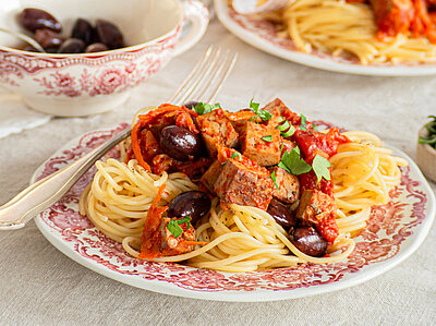 Rote Tofuwürfel mit rotem Gemüse und Oliven auf Nudeln auf 2 Tellern mit Tischdekoration.