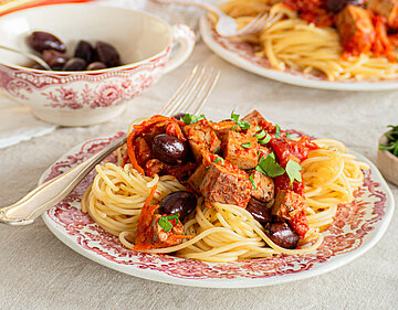 Rote Tofuwürfel mit rotem Gemüse und Oliven auf Nudeln auf 2 Tellern mit Tischdekoration.