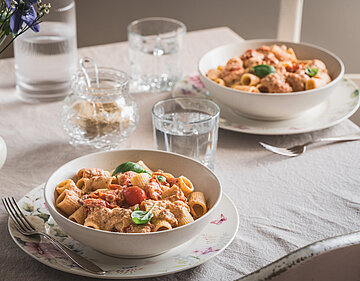 Pasta mit Soße, Tomaten und Basilikum auf Tellern und Wassergläser auf einem Tischtuch mit Dekoration.