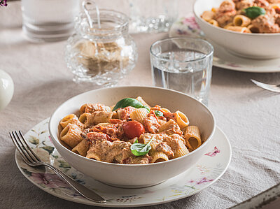 Pasta mit Soße, Tomaten und Basilikum auf Tellern und Wassergläser auf einem Tischtuch mit Dekoration.