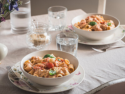 Pasta mit Soße, Tomaten und Basilikum auf Tellern und Wassergläser auf einem Tischtuch mit Dekoration.