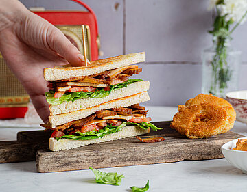 Zwei Toastscheiben belegt mit Bacon auf einem Holzbrett und Dekoration wird von einer Hand gehalten.