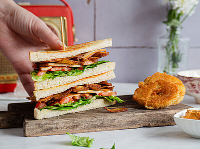 Zwei Toastscheiben belegt mit Bacon auf einem Holzbrett und Dekoration wird von einer Hand gehalten.