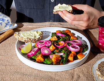 Tofuwürfel mit grünem Gemüse und Zwiebelringen auf einem Teller, dazu Brot mit hellem Aufstrich.