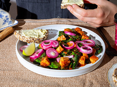 Tofuwürfel mit grünem Gemüse und Zwiebelringen auf einem Teller, dazu Brot mit hellem Aufstrich.