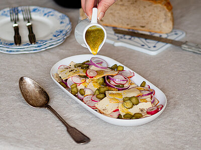Dünne Scheiben Tofu mit Zwiebeln, Gurken und Radieschen in einer Schale, jemand gießt gelben Dressing darauf, mit Tischdekoration.