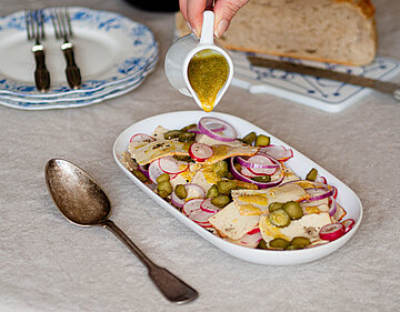 Dünne Scheiben Tofu mit Zwiebeln, Gurken und Radieschen in einer Schale, jemand gießt gelben Dressing darauf, mit Tischdekoration.