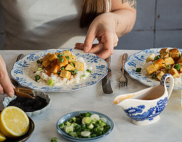 Tofu süß-sauer schnell einfach Jemand gießt Soße über Tofuwürfel auf Reis und Lauch auf einem Teller mit Tischdekoration.