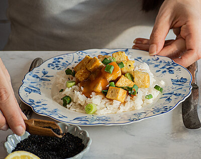 Jemand gießt Soße über Tofuwürfel auf Reis und Lauch auf einem Teller mit Tischdekoration.