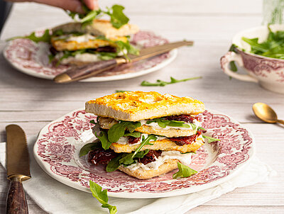 Vier übereinanderliegende Waffeln, dazwischen Tofu, getrocktete Tomaten und Salat aufeinem Teller mit Tischdekoration.