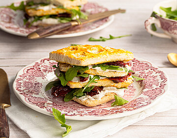 Vier übereinanderliegende Waffeln, dazwischen Tofu, getrocktete Tomaten und Salat aufeinem Teller mit Tischdekoration.