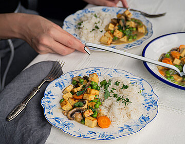 Tofustückchen, Pilze, Möhren und Erbsen an Reis auf einem Teller mit Tischdekoration.