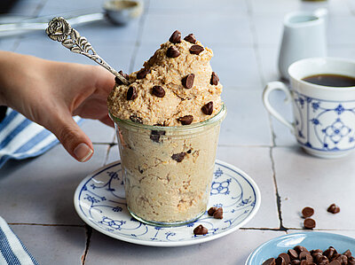 Hellbraune Creme mit Schokostückchen in einem Glas, das von einer Hand gehalten wird, auf einem Teller mit Tischdekoration.