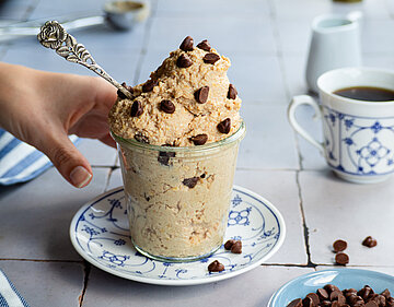 Hellbraune Creme mit Schokostückchen in einem Glas, das von einer Hand gehalten wird, auf einem Teller mit Tischdekoration.
