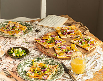 Vlammkuchen auf 2 Tellern und einem Holzbrett mit Zweibeln, Tofu und Lauch mit Tischdekoration.