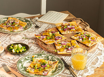 Vlammkuchen auf 2 Tellern und einem Holzbrett mit Zweibeln, Tofu und Lauch mit Tischdekoration.