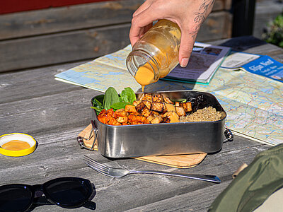 Tofu-Würfel mit Quinoa und Salat in einer Metall-Schüssel, jemand gießt eine gelbe Soße darauf auf einem Holztisch.