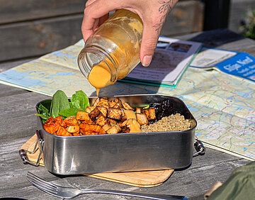 Quinoa-Tofu-Bowl Tofu-Würfel mit Quinoa und Salat in einer Metall-Schüssel, jemand gießt eine gelbe Soße darauf auf einem Holztisch.