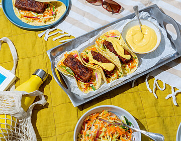 Tacos mit gebratenen Tofuscheiben mit buntem Salat und gelber Sauce auf einem Brett mit Tischdekoration.