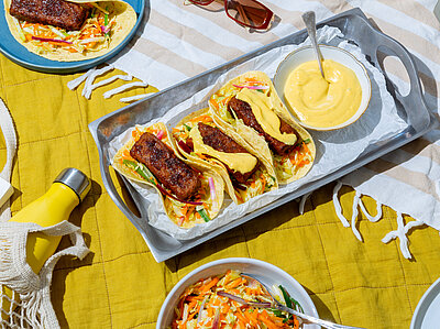 Tacos mit gebratenen Tofuscheiben mit buntem Salat und gelber Sauce auf einem Brett mit Tischdekoration.