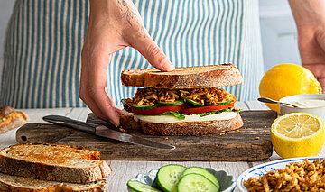 Zwischen 2 Graubrotscheiben Tofu-Stückchen und Salat auf einem Holzbrett und Dekoration.