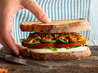 Zwischen 2 Graubrotscheiben Tofu-Stückchen und Salat auf einem Holzbrett und Dekoration.