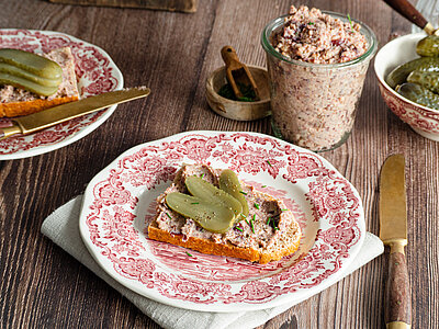 Ein Glas mit einer Tofupaste, daneben ein Brot auf einem Teller mit der Tofupaste und Gurkenscheiben mit Dekoration.