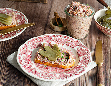 Ein Glas mit einer Tofupaste, daneben ein Brot auf einem Teller mit der Tofupaste und Gurkenscheiben mit Dekoration.