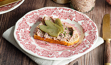 Ein Glas mit einer Tofupaste, daneben ein Brot auf einem Teller mit der Tofupaste und Gurkenscheiben mit Dekoration.